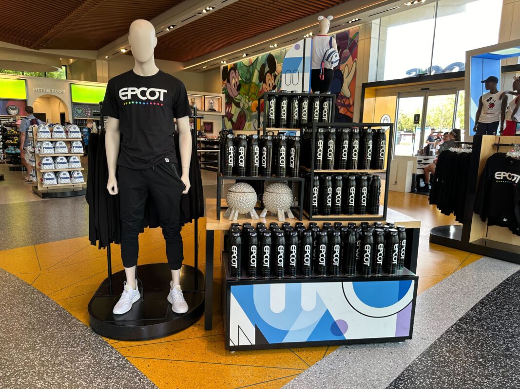 Display of EPCOT merchandise with a mannequin wearing a black EPCOT shirt standing next to shelves filled with EPCOT water bottles and decorative items in a store.