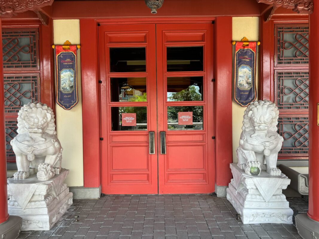 A pair of stone lion statues flank a set of closed red double doors at the entrance of a Chinese-style building, with intricate designs and decorative signs above the doors—a scene that feels like it's been taken straight from an architectural guide rather than an auto draft.