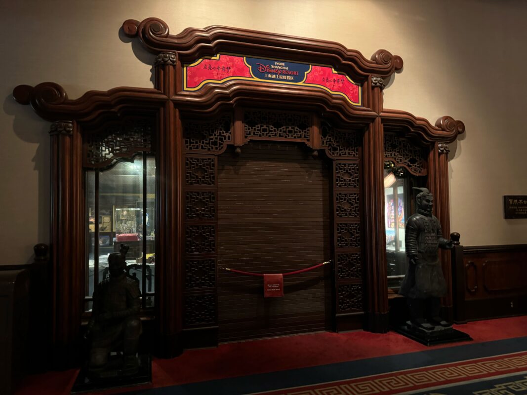 Dimly lit hallway with a closed ornate wooden door, flanked by two statues, one seated and one standing. A sign above indicates "Shanghai Disneyland" in English and Chinese. A red rope is in front, lending an air of exclusivity that feels almost like the prelude to an auto draft of adventure.