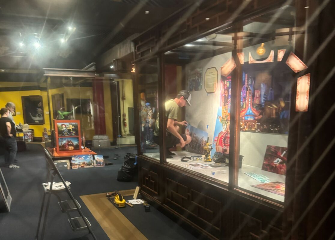 A person works on setting up a display inside a museum exhibit; tools and materials are scattered on the floor, and a partially constructed diorama is visible beside an auto draft blueprint.