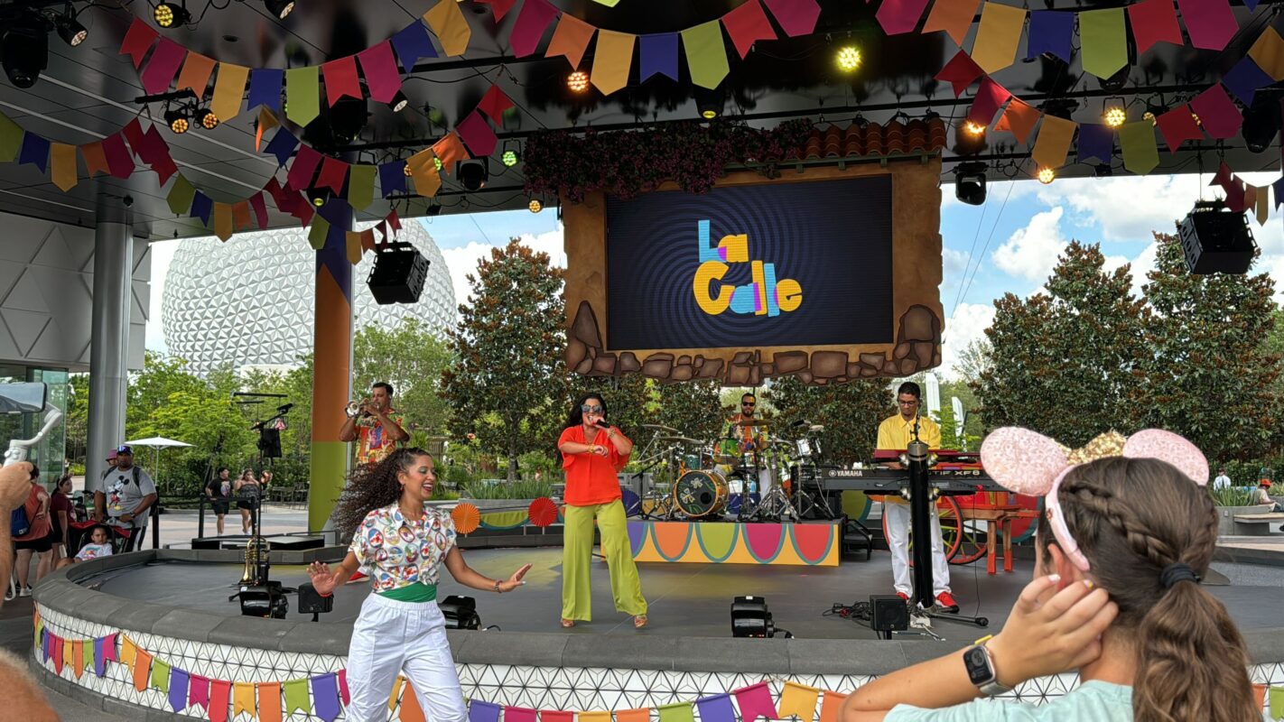 La Calle Band performs on a decorated outdoor stage with colorful banners at EPCOT's CommuniCore Plaza. A female dancer engages with the audience in the foreground, while onlookers, including a person with mouse ears, watch.