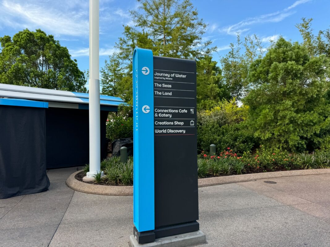 Signpost in a park with directional information for attractions including Journey of Water, The Seas, The Land, Connections Cafe & Eatery, Creations Shop, and World Discovery. Trees and building in background.