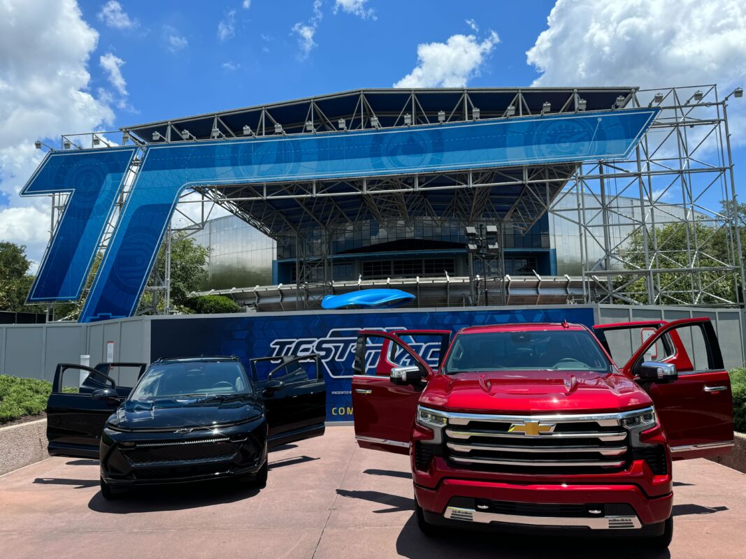 Two Chevrolet vehicles, one black and one red, are parked in front of a large blue and metal building adorned with the 'Test Track' sign. The sky is partially cloudy, adding to the atmosphere of the auto draft setting.