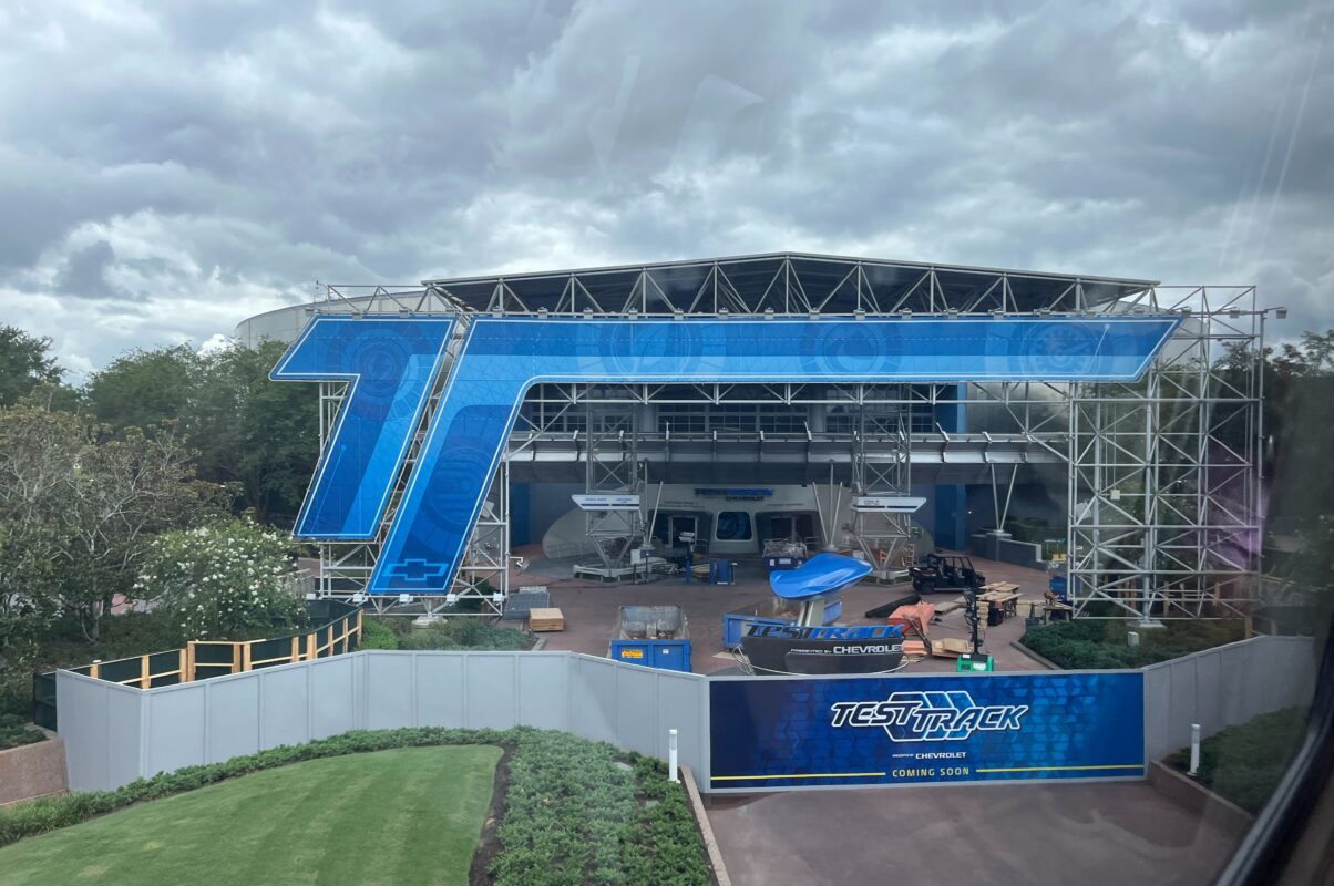 View of the Test Track attraction under construction with scaffolding and construction materials visible. Large blue signage with "T" and "Test Track" branding is displayed prominently.