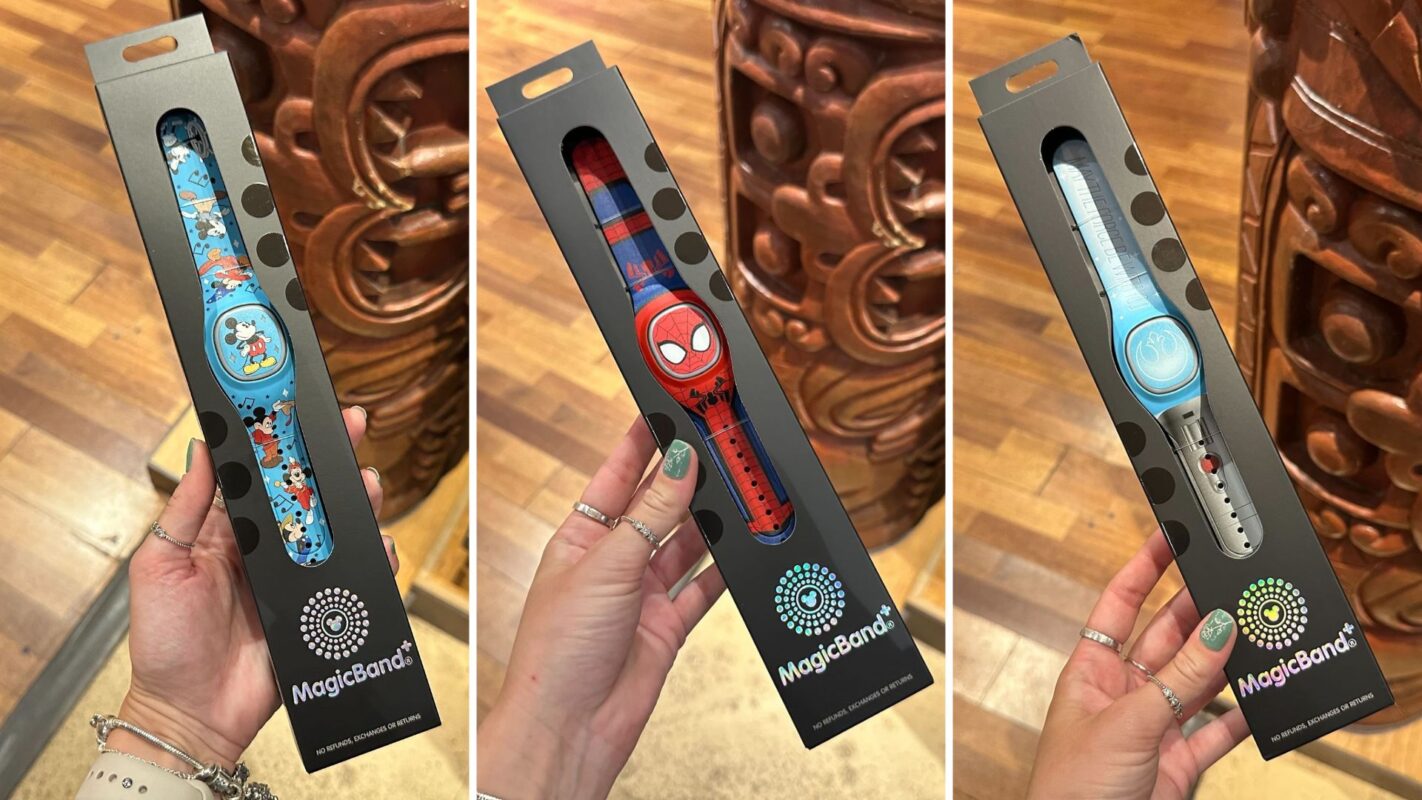 Three MagicBand+ boxes held by hands, featuring different bands: blue with characters, red with a superhero, and blue and white with design elements. Wooden floor and decor in the background.