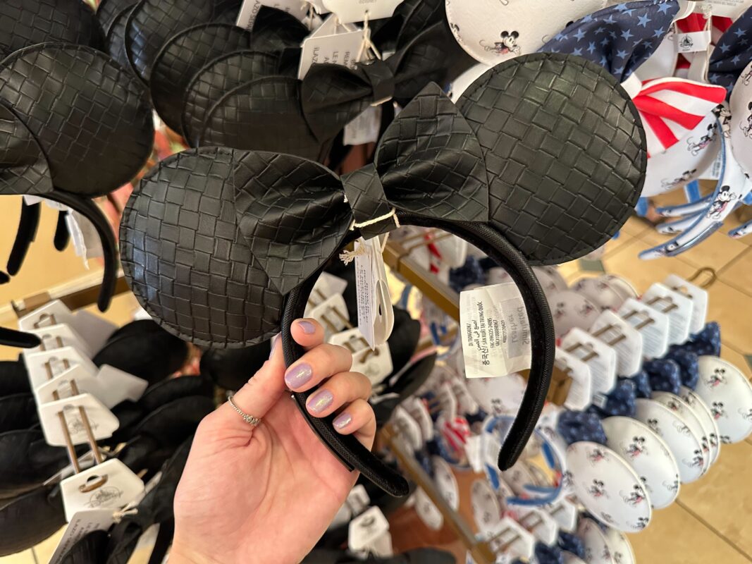 A hand holds a black, woven texture mouse ears headband displayed among similar headbands on a store rack.