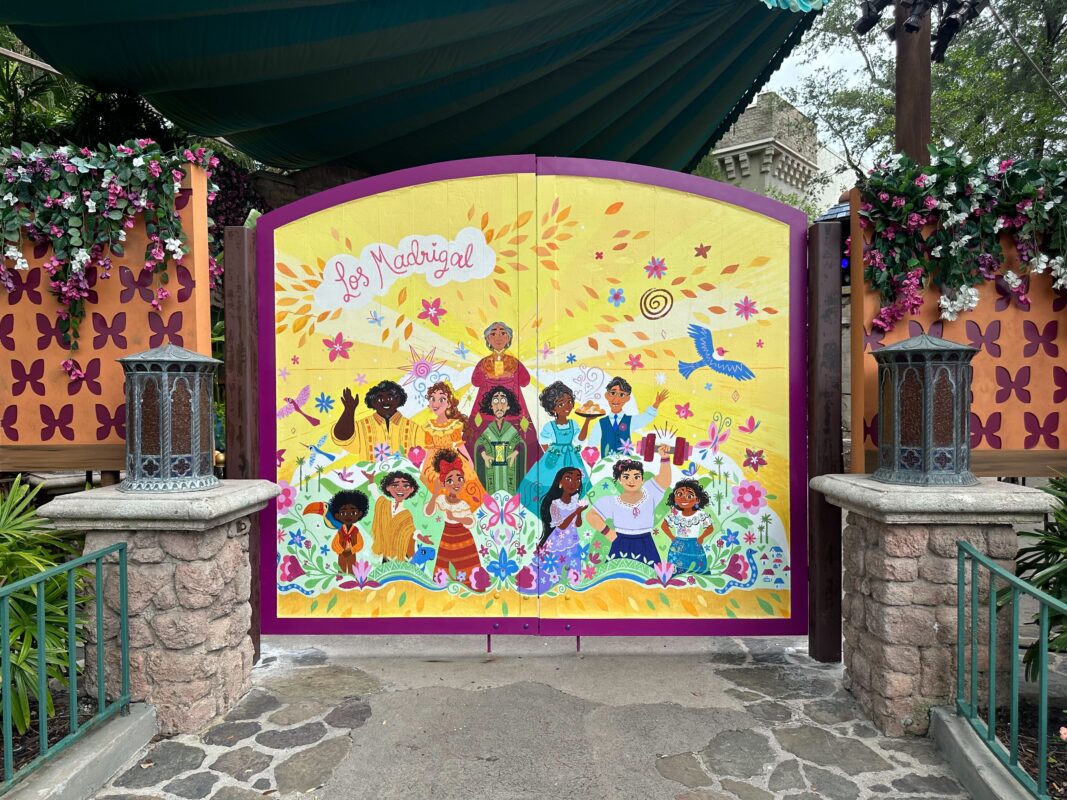 A vibrant mural on a gate features a diverse group of characters from Disney's "Encanto," labeled "Los Madrigal." The gate, perfect for a Mirabel meet and greet, is adorned with flowers and surrounded by decorative lanterns and foliage.