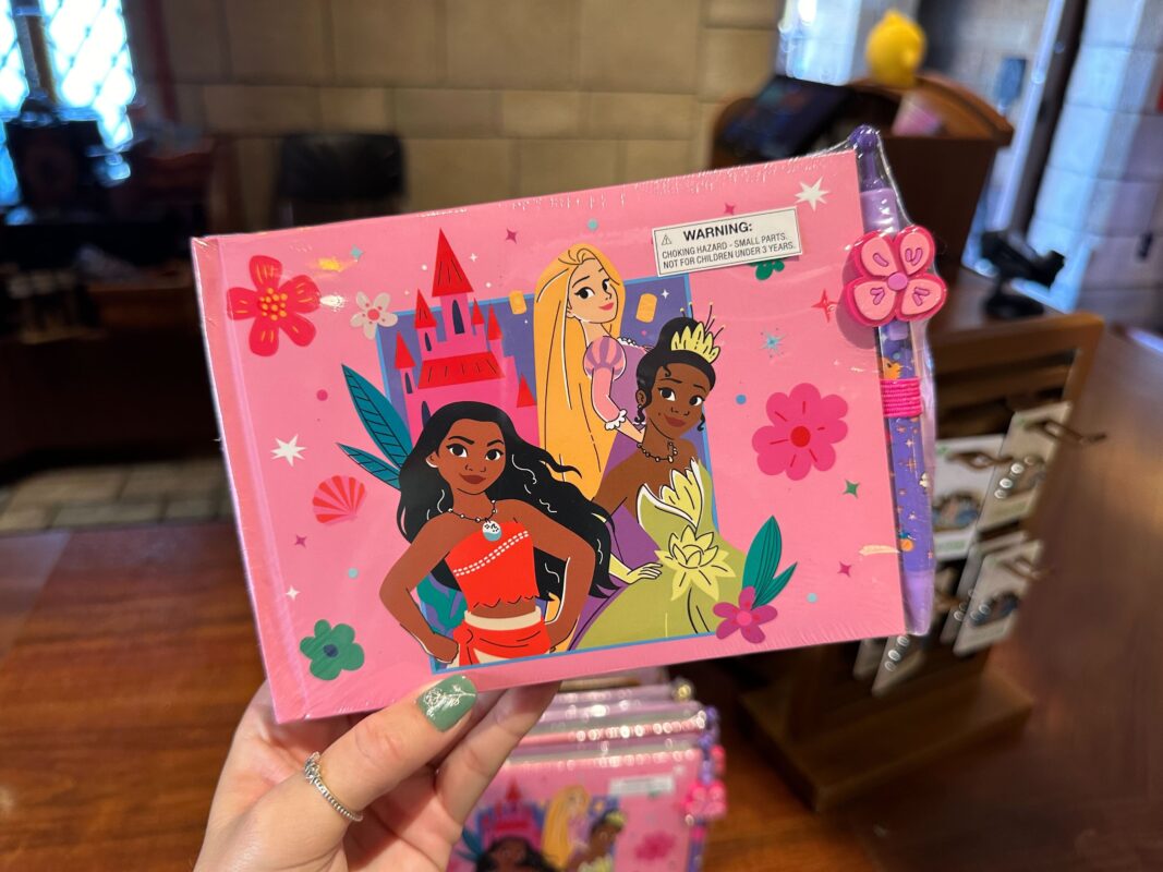 A photo of a person holding a pink notebook featuring animated characters against a colorful background, with a warning sticker and a pen attached on the side.