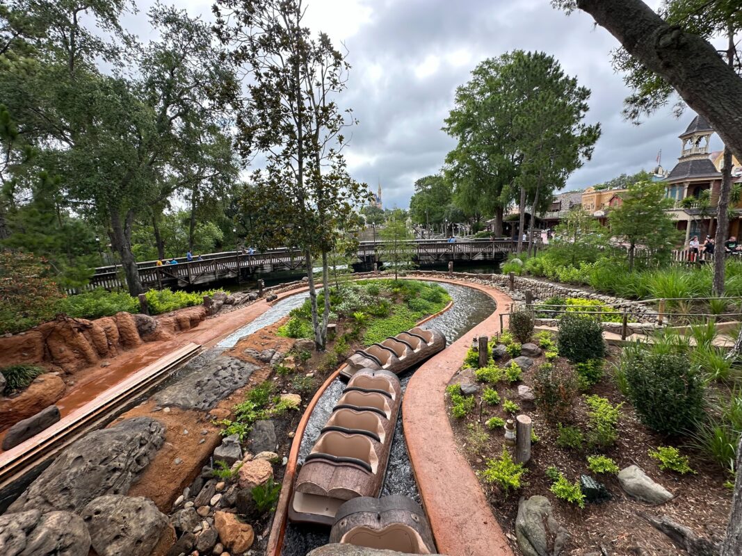 An outdoor amusement park ride, Tiana's Bayou Adventure, features empty log boats on a water track, surrounded by green trees, various plants, and rocky landscaping under a clouded sky. Currently not yet operating but soon available for Passholder Previews.