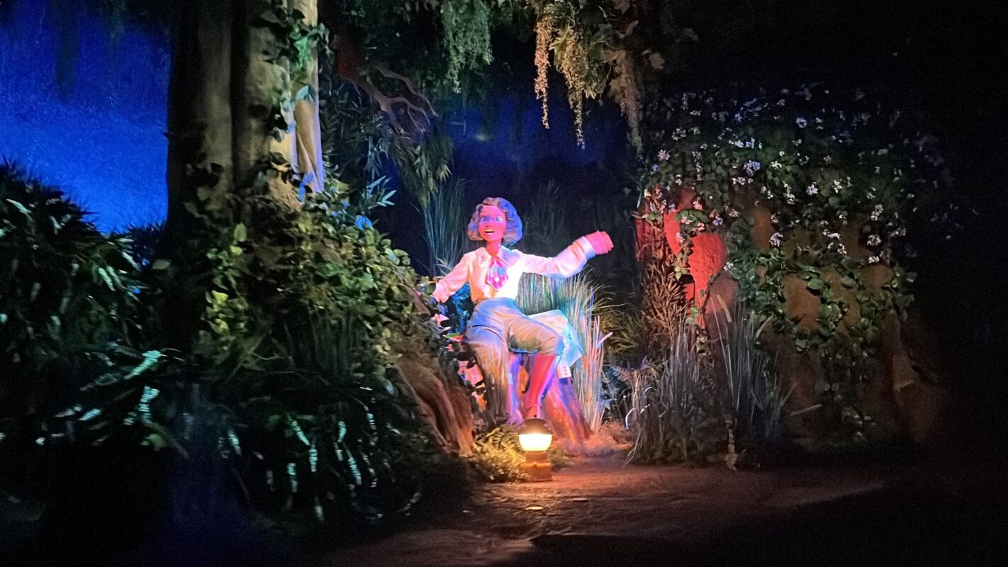 A Tiana animatronic seated amidst lush foliage in a dimly lit bayou setting with some flowers visible, illuminated by a spotlight.