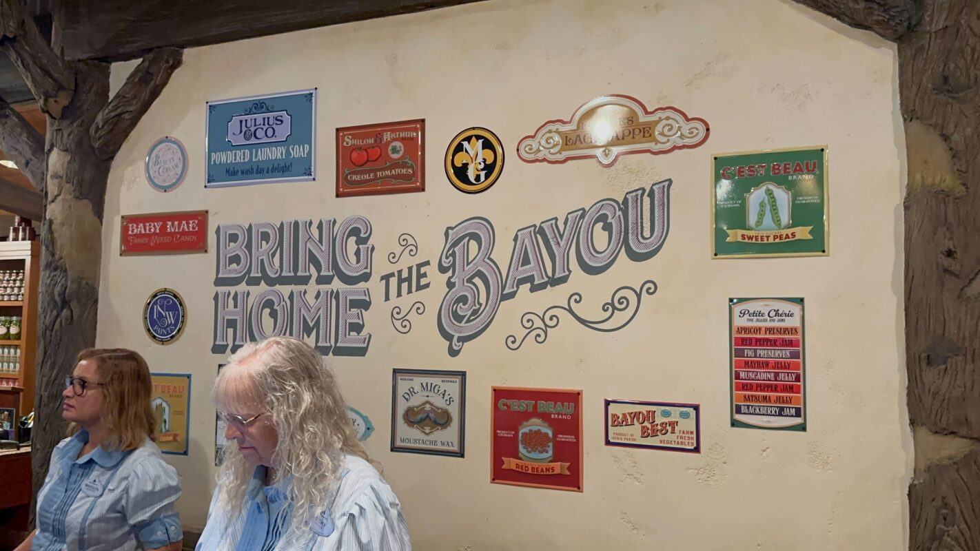 Two people stand in front of a wall adorned with various vintage-style signs, including messages like "Bring Home the Bayou" and "Jolie's Co." The atmosphere is reminiscent of a southern bayou-themed setting.