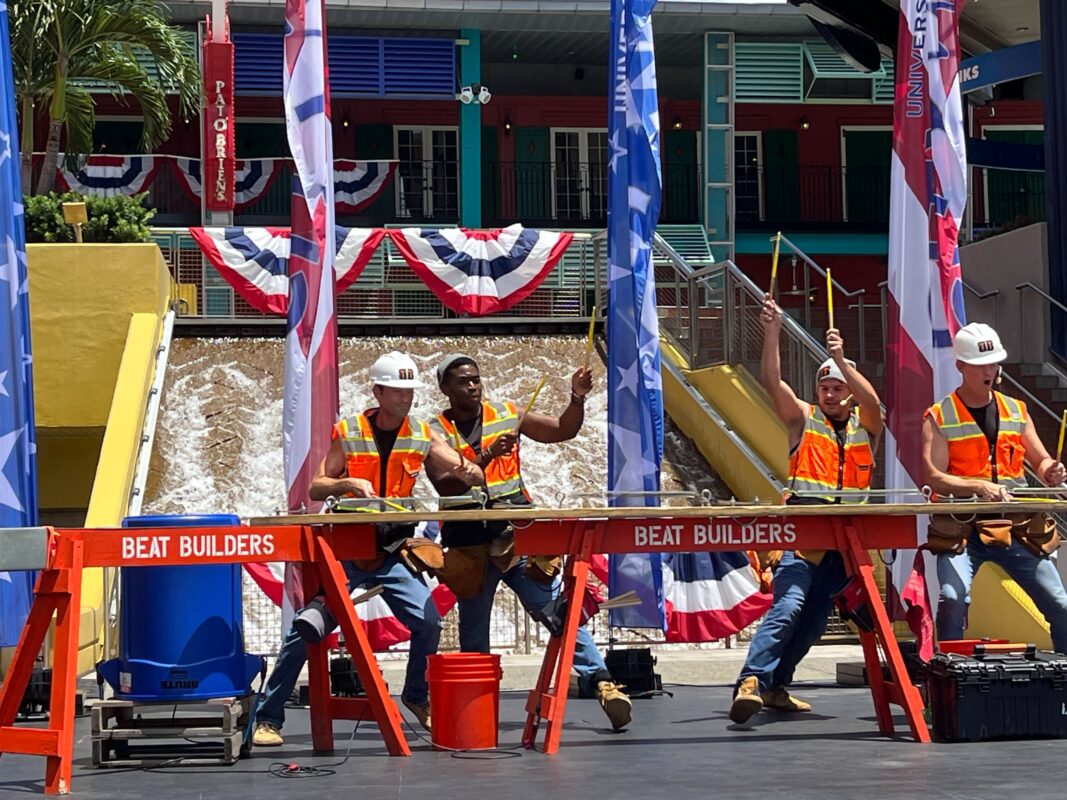 Four construction workers in orange safety vests and hard hats are performing on a stage set with decked beams labeled "Beat Builders." American flags and bunting decorate the background.