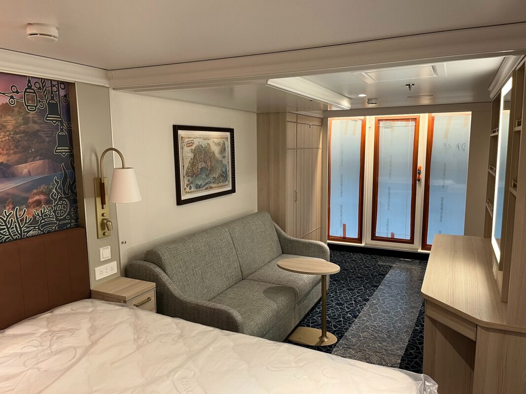 A cozy Disney Cruise Ship cabin with a bed, a grey sofa, a small round table, and a balcony door with frosted glass panels. The walls feature a map and an artwork. This lovely space is one of the charming Disney Treasure Staterooms awaiting your final installation of comfort items.