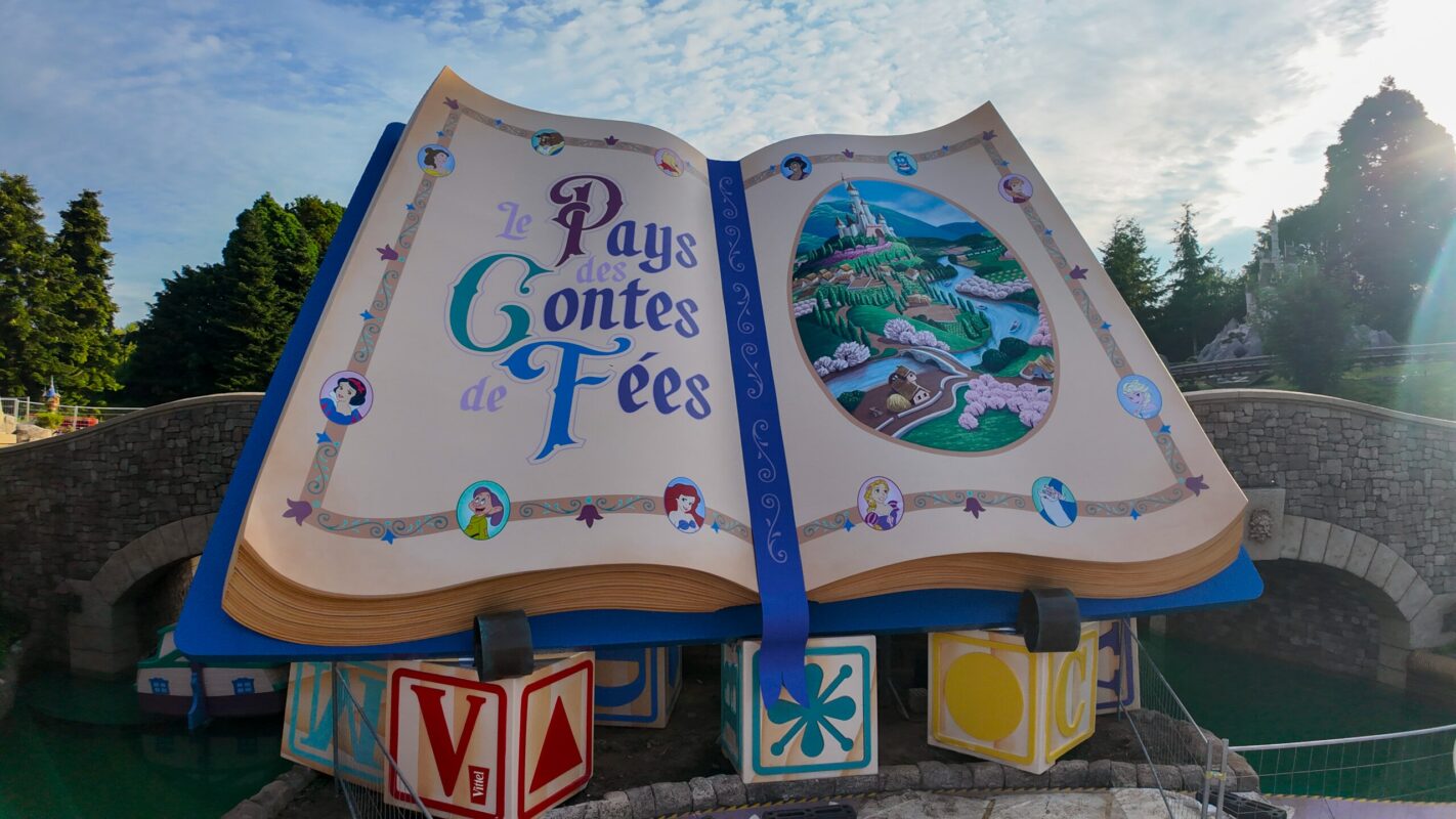 An open storybook sculpture with illustrated pages titled "Le Pays des Contes de Fées" stands in front of a stone bridge surrounded by trees under a partly cloudy sky.