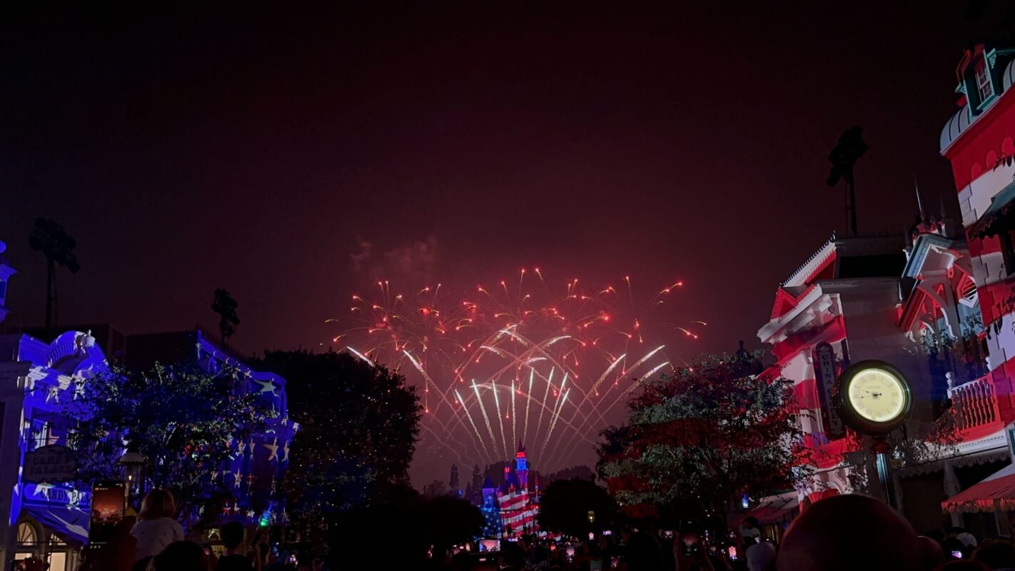 Nighttime fireworks display over a theme park, with buildings and trees illuminated in red, white, and blue lights. A crowd of people watches the spectacle.
