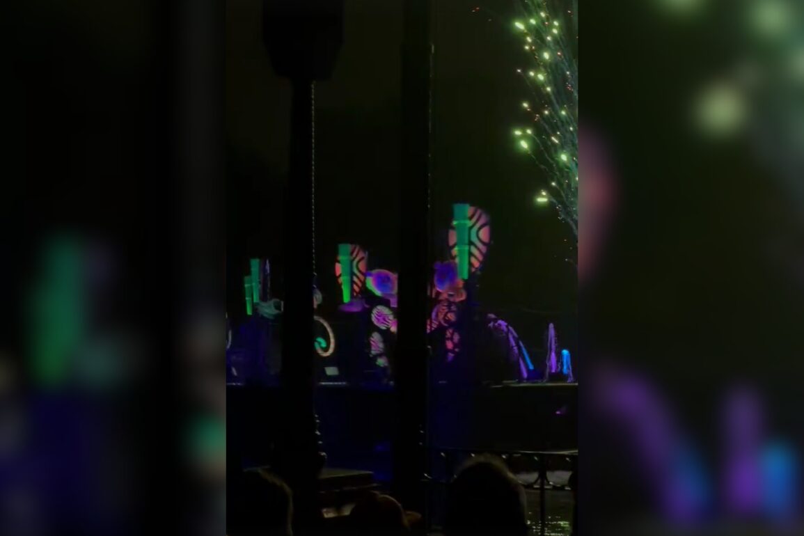 A nighttime show with illuminated, colorful abstract shapes and patterns projected onto stage structures with fireworks in the background.