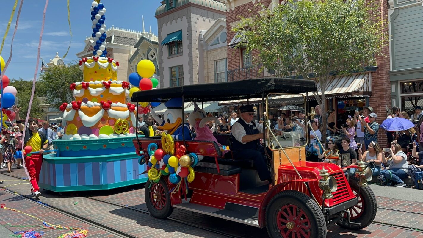 A red vintage car and a colorful cake-themed parade float travel down a street lined with spectators during the 69th Anniversary Cavalcade. A performer holds streamers near the float decorated with balloons and a character in blue and yellow attire.