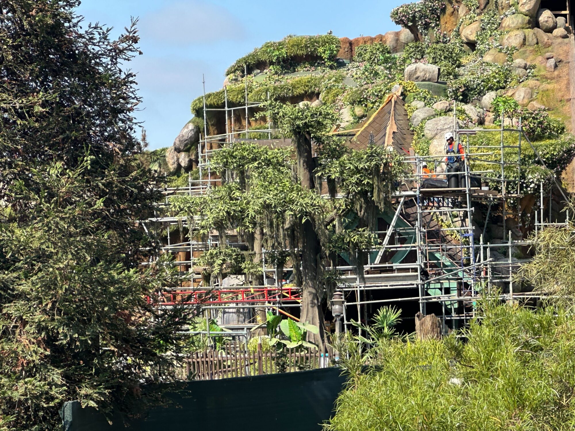 Construction site with scaffolding around a large, tree-covered rock formation. Workers are present, and various building materials and equipment are visible. Trees and plants surround the area.