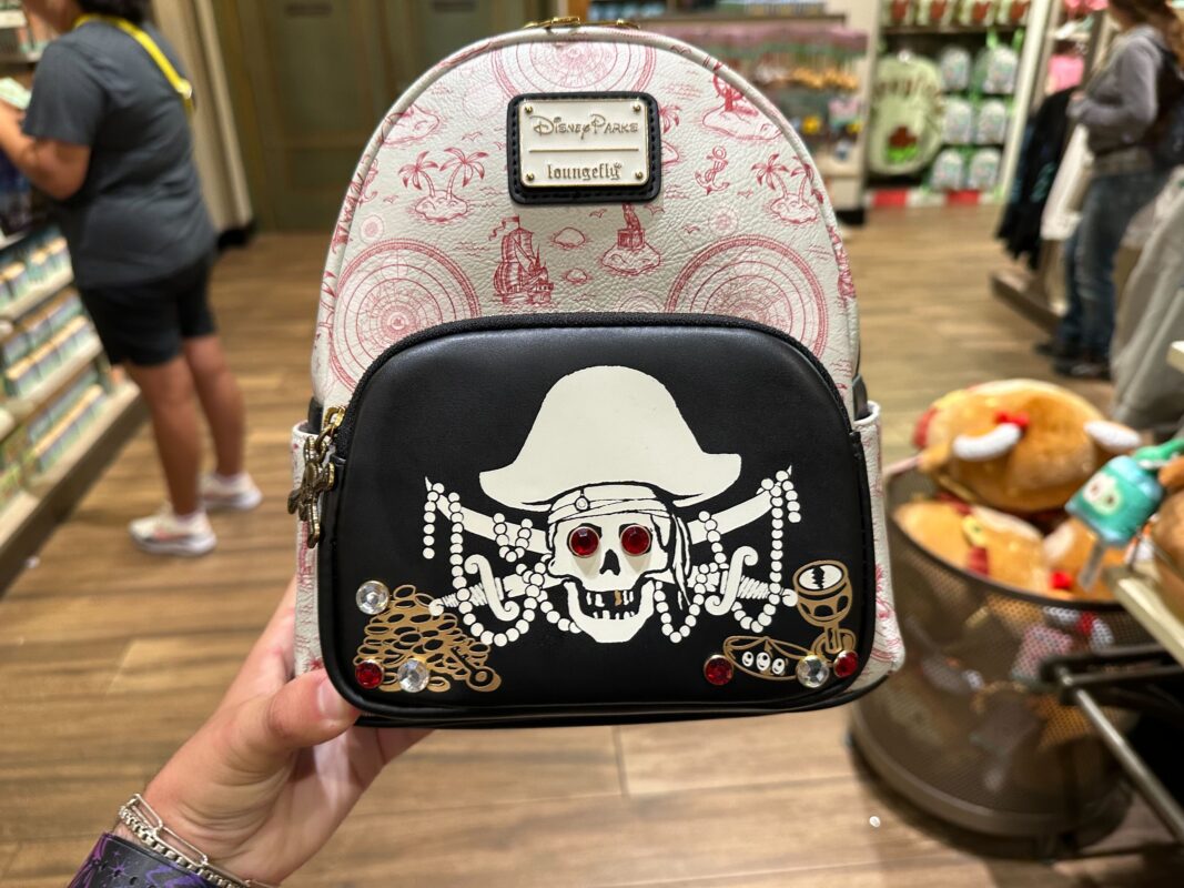 A hand holding a small backpack with a pirate skull and crossbones design. The skull has red eyes and is wearing a pirate hat. The background features various items in a store setting.