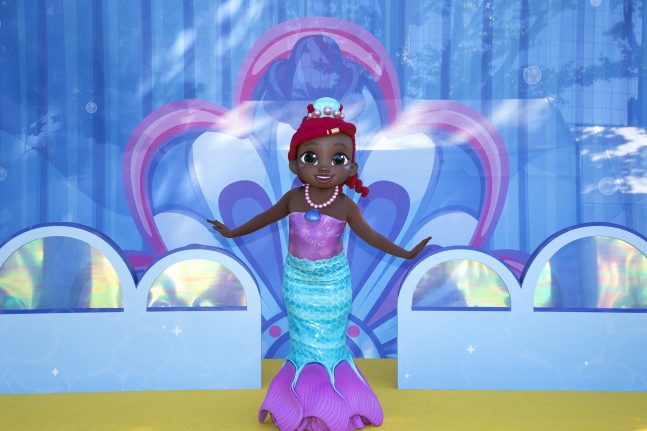 A doll with dark skin and dark hair is dressed as a mermaid with a pink top and blue tail, in front of a colorful underwater-themed backdrop.