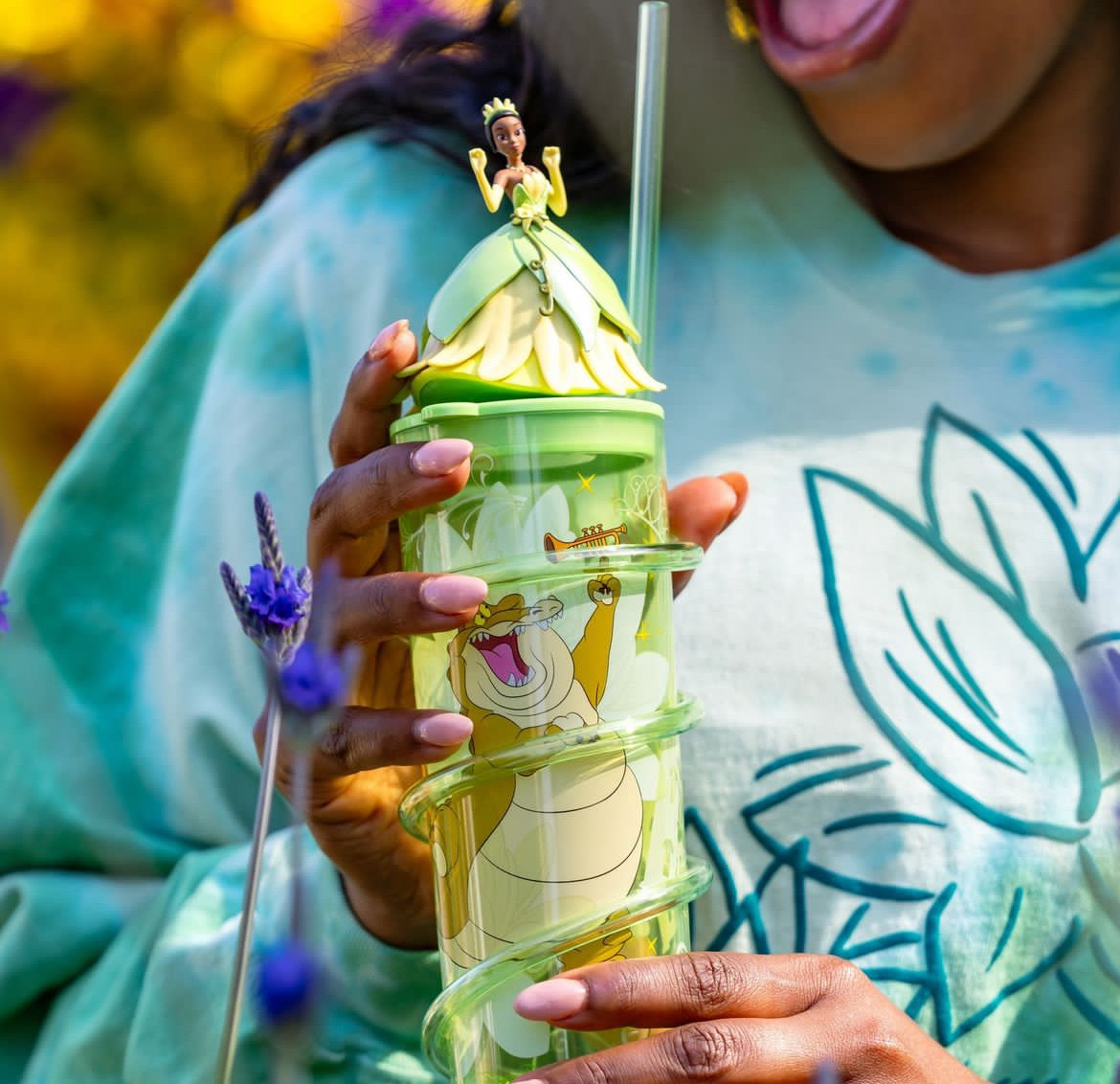 A person holds a green spiral-shaped Princess Tiana Sipper with a princess figurine lid and straw. The cup also features an animated bear character. They are wearing a green Disneyland Resort shirt with a floral graphic.