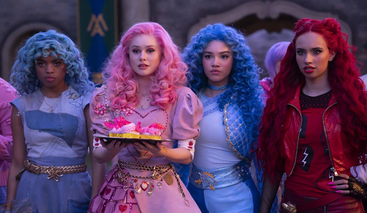 Four young women with colorful hair and outfits stand together, including a pink-haired woman holding a tray of cupcakes, reminiscent of the vibrant characters from Disney+'s "Descendants: The Rise of the Red," which shattered viewership records.