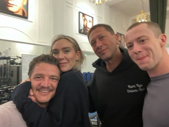 Four people are indoors, facing the camera. One individual is hugging another from the side, and all are smiling like a fantastic four. There are framed pictures on the wall behind them.