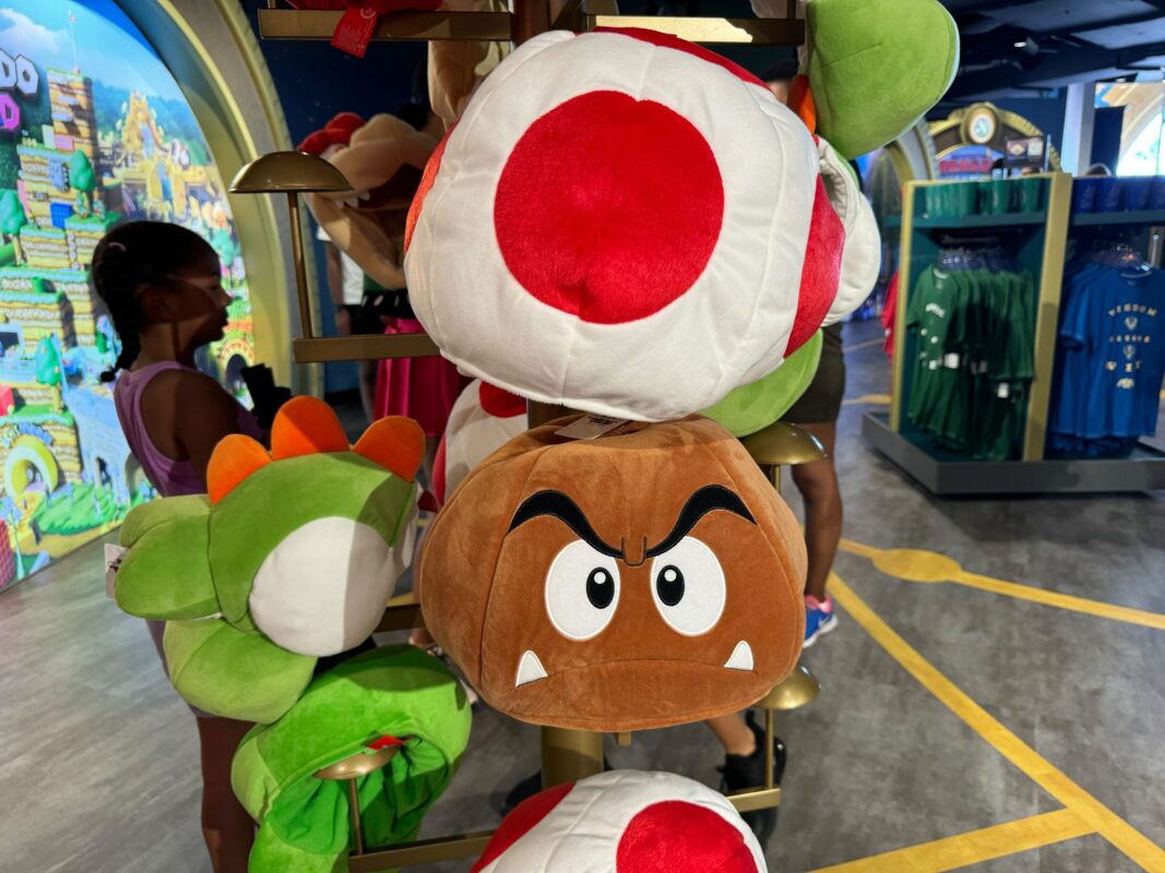 A display of plush toys, including a green and white dinosaur, a red and white mushroom, and a brown character with angry eyes and fangs, inside a store at Universal Orlando Resort with two individuals in Super Nintendo World-themed hats in the background.