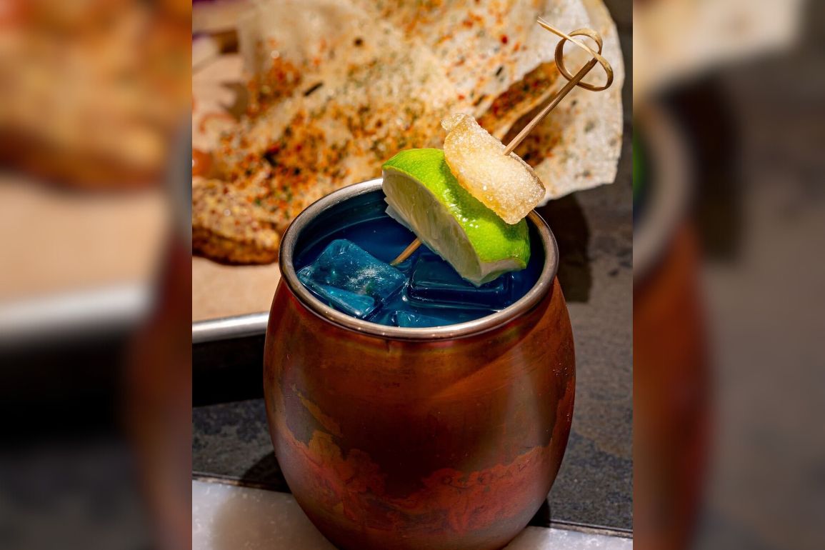 A copper cup filled with blue liquid and ice cubes, garnished with a lime wedge and a sugar cube on a skewer, holds the mysterious Krill Elixir. A toasted piece of bread with seasoning is in the background.