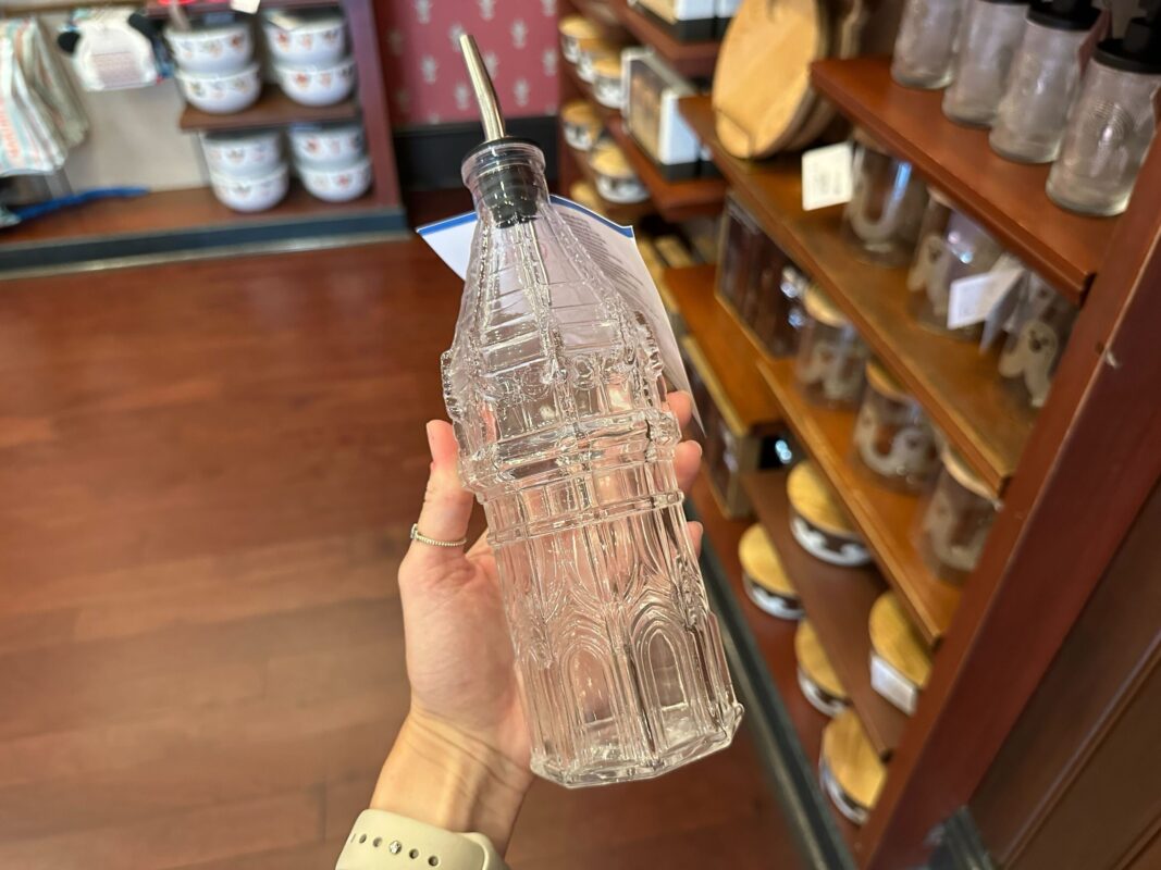 A hand holds a clear, intricately designed glass bottle with a metal spout. In the background, shelves display various kitchen items.