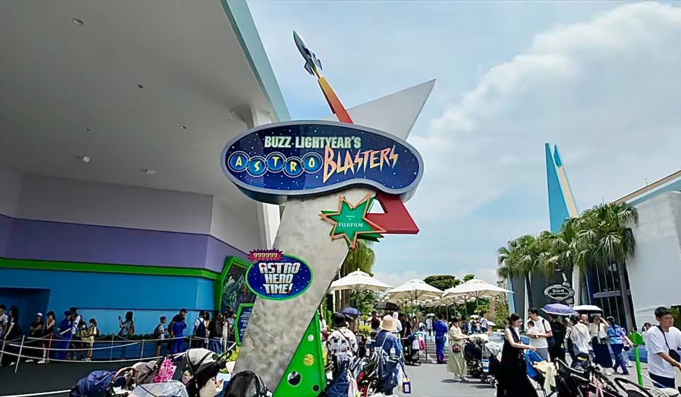 Entrance sign for Buzz Lightyear's Astro Blasters attraction at a theme park. The area is crowded with people waiting in line, several strollers parked nearby, and excited visitors eager to experience the adventure inspired by the Full Ride Video.