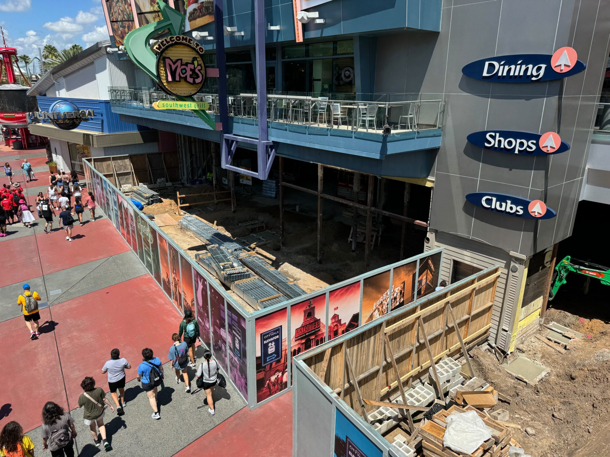 A view of a construction site behind barricades in an outdoor shopping area at CityWalk, with signs for Dining, Shops, and Clubs visible on a nearby building. People are walking along the pathways.