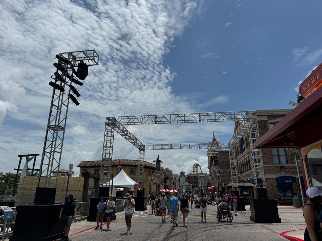 People walk along a street lined with buildings and under several metal truss structures on a sunny day with a partly cloudy sky.