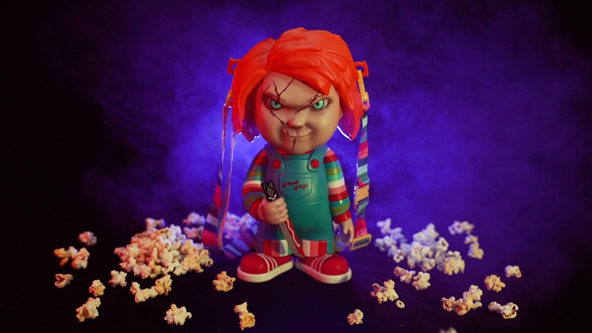 A Chucky doll with orange hair, scars on its face, and colorful clothes stands menacingly amid scattered popcorn with a smoky purple background, perfect for Halloween Horror Nights.