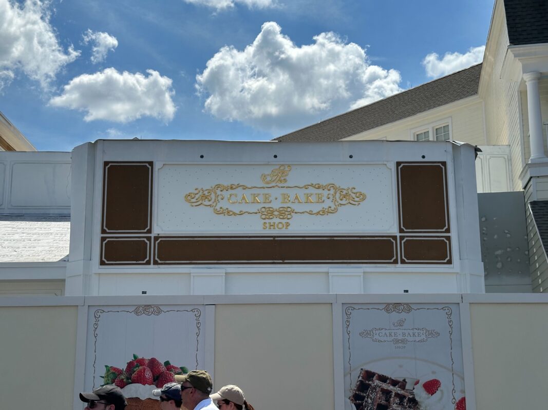 The photo shows the facade of "The Cake Bake Shop" with a decorative sign, against a partly cloudy sky. Several people stand in front of a barrier below the sign, seemingly captivated by the impeccable design that looks almost auto draft perfect.