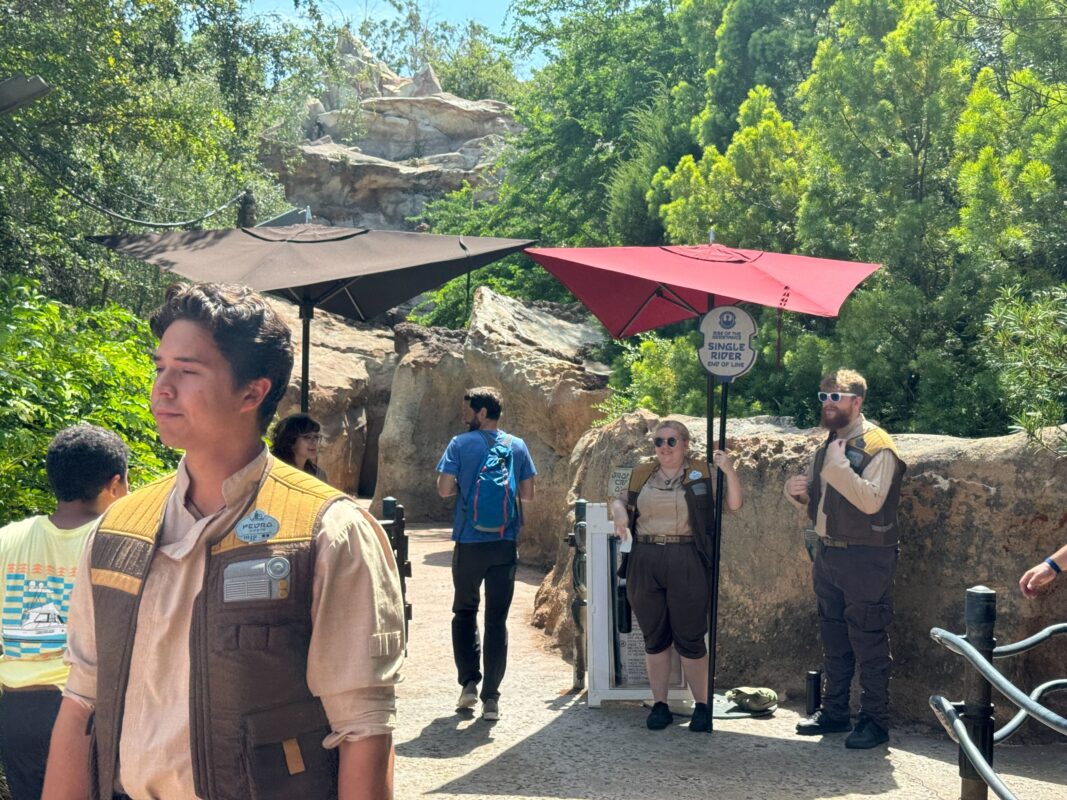Several park staff in themed attire stand near an area with umbrellas and a sign indicating a "Single Rider Line" for Rise of the Resistance, while visitors walk in the background.