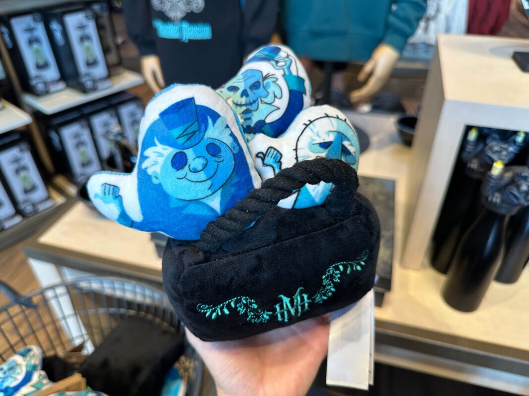 A person holding a small plush toy depicting three blue characters emerging from a black container with decorative green embroidery. The background shows a store display with other Haunted Mansion merchandise at Disney World. You may need to ask about prices for these unique collectibles!