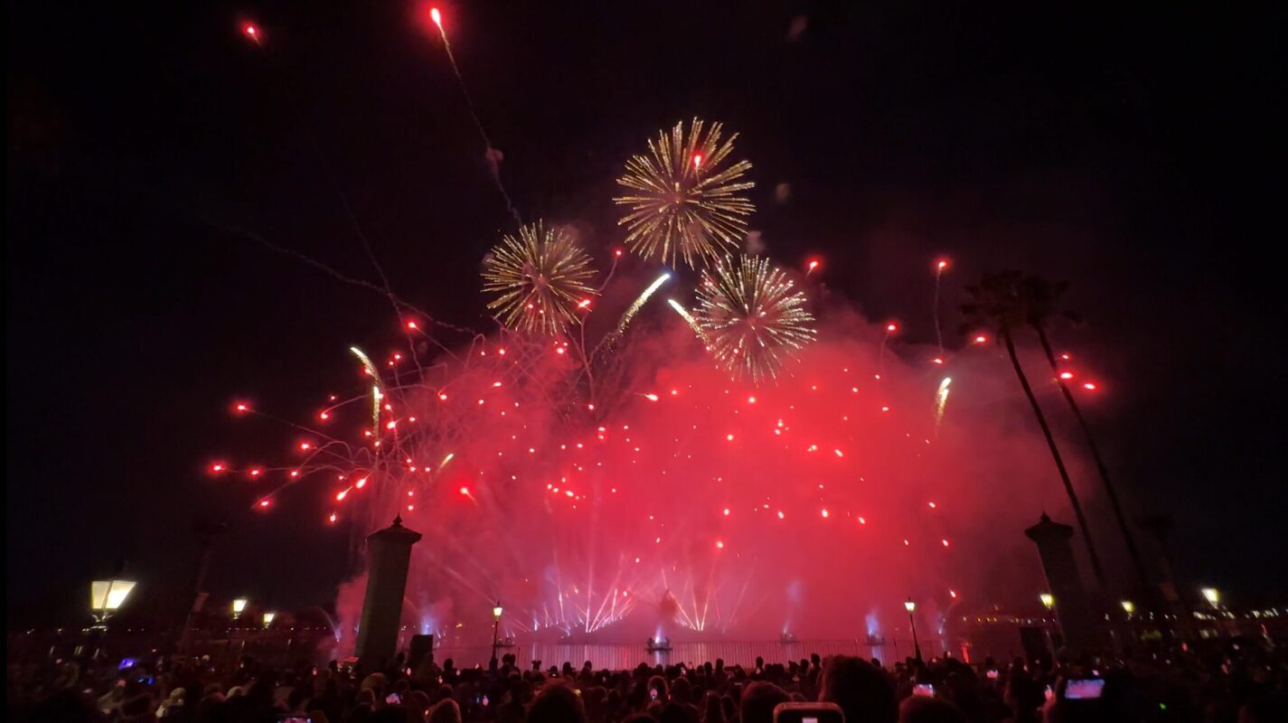 A large crowd watches a vibrant Fourth of July fireworks display in the night sky over EPCOT. Bright red and yellow fireworks light up the area, with some palm trees visible on the right side.