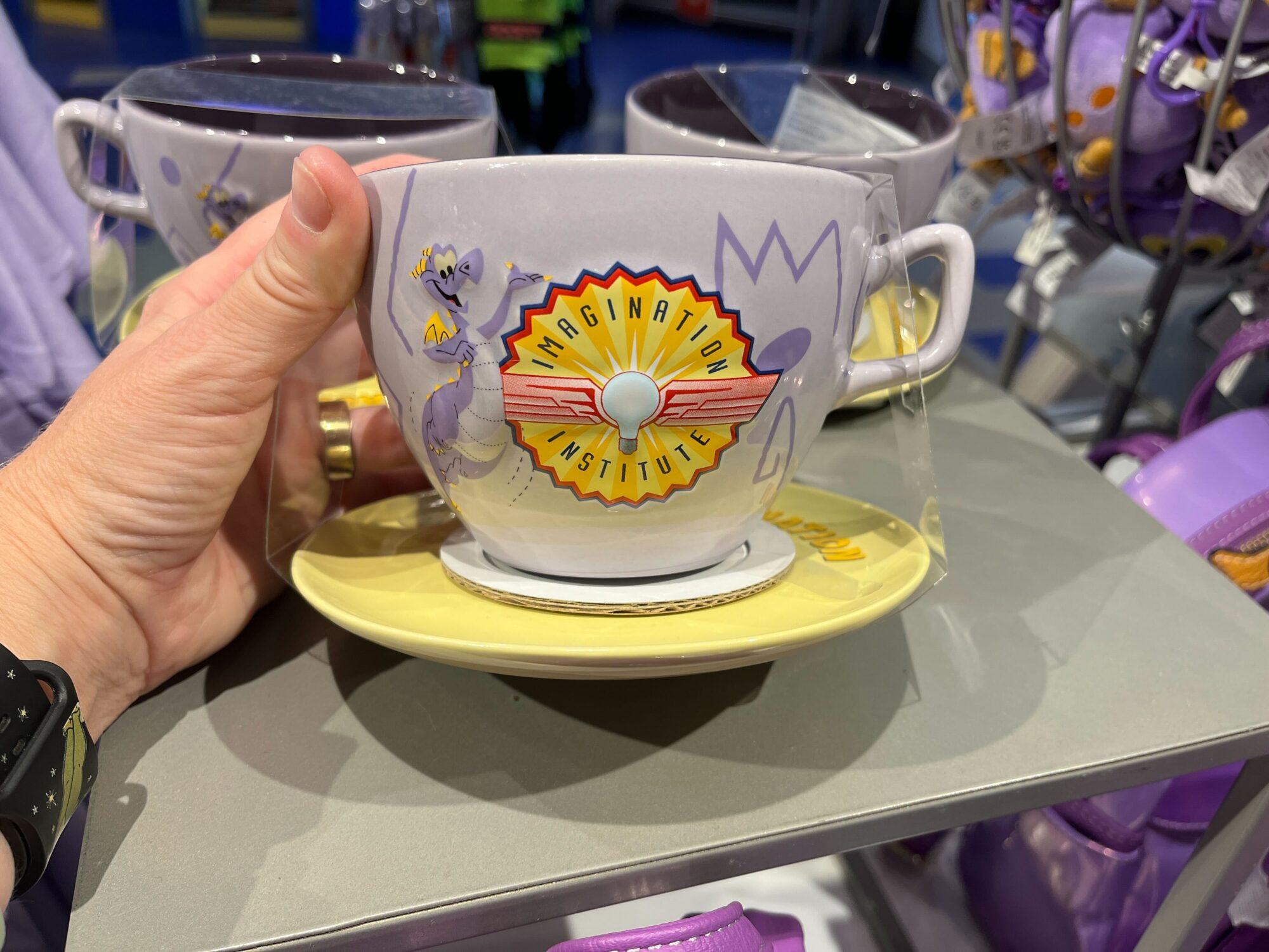 A person holds a large Figment Mug with the "Imagination Institute" logo and a purple cartoon character, displayed among similar cups in an EPCOT store.