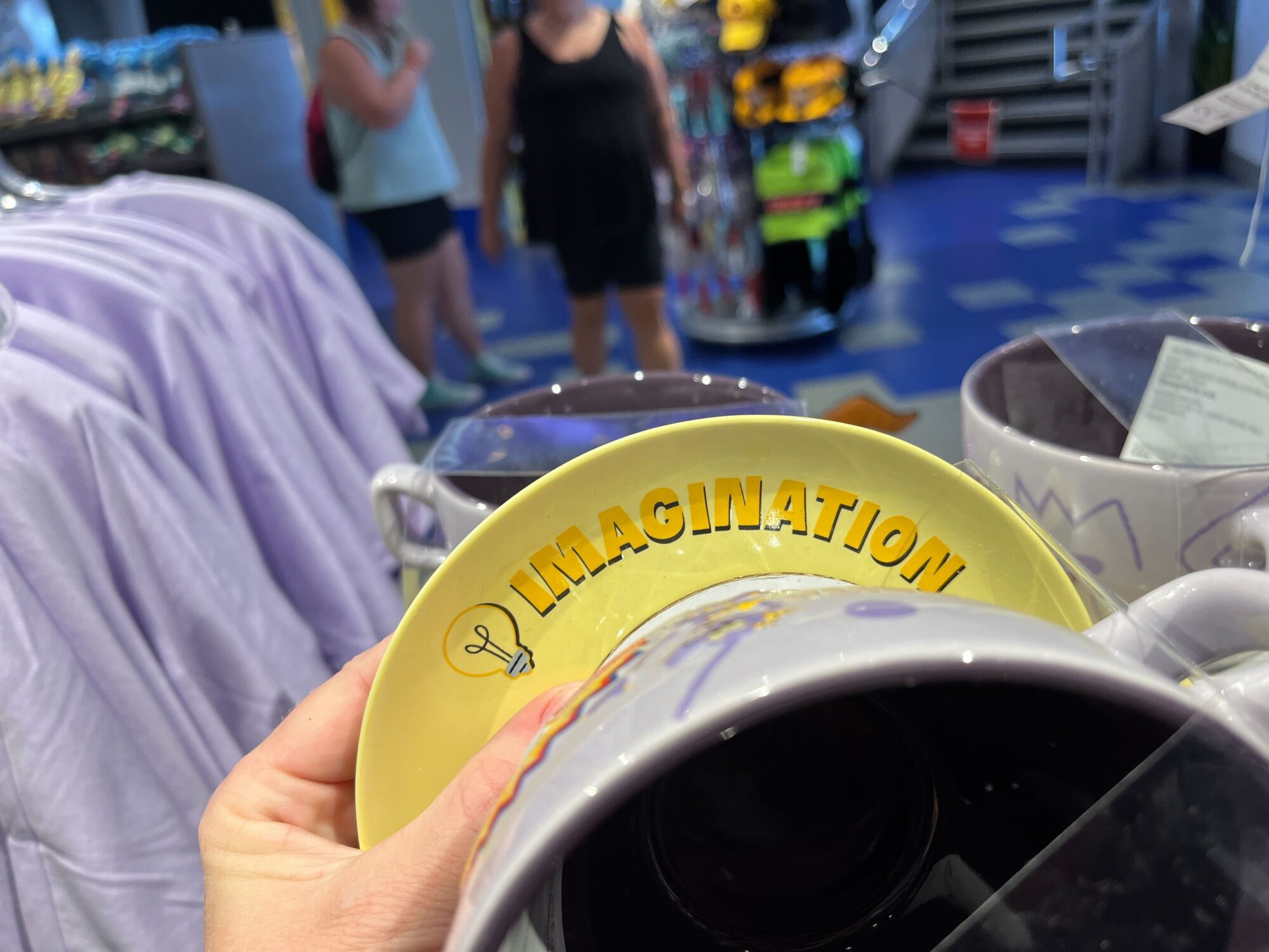 A hand holding a yellow Figment mug with the word "IMAGINATION" and a lightbulb icon on the rim. The background shows a store with people, clothing, and other items for sale—evoking the creative spirit of EPCOT.