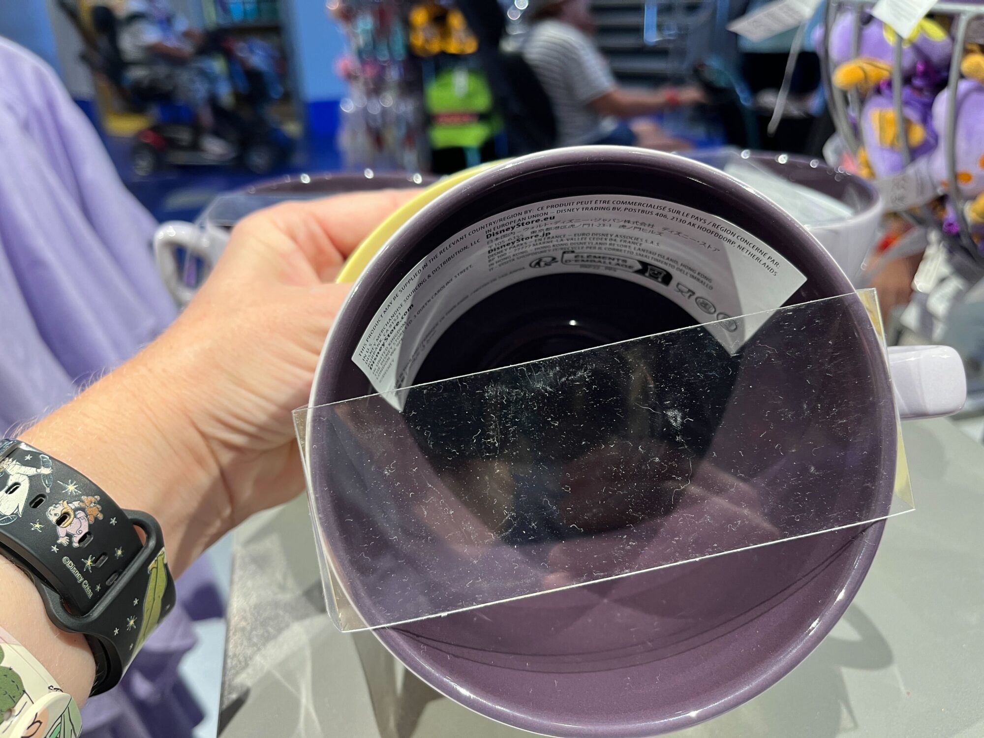 A person holds a large purple Figment Mug with a barcode and price tag inside. The background shows various colorful items on display in an EPCOT store setting.