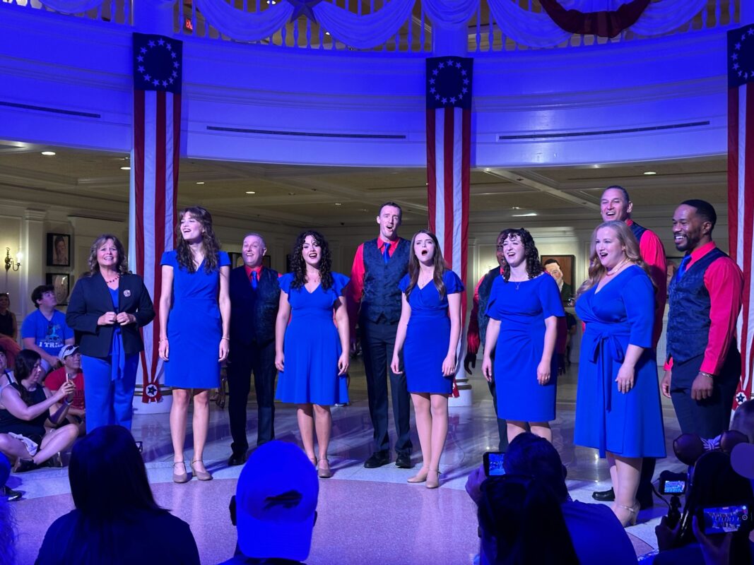 A group of men and women in blue and red outfits sing on a stage with American-themed decor, including flags and white drapery. Spectators watch from the foreground.