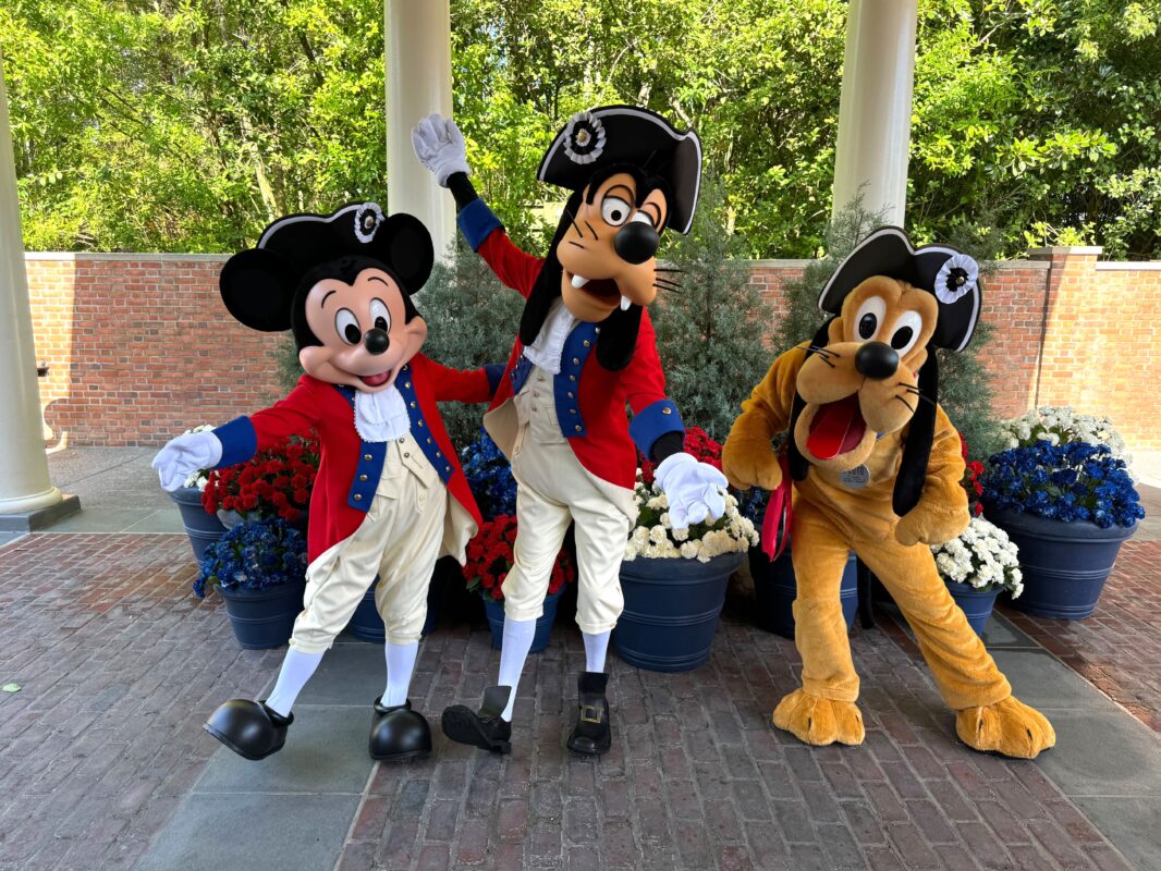 Three costumed characters dressed in colonial-style outfits stand in front of potted plants and a brick wall, embodying the spirit of The American Adventure.