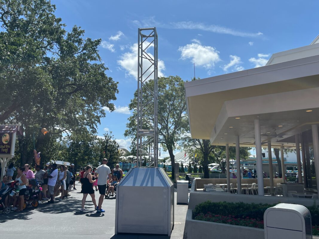 People walking and sitting near a white building with an outdoor seating area, surrounded by trees on a sunny day. There is a tall metal structure next to the building.