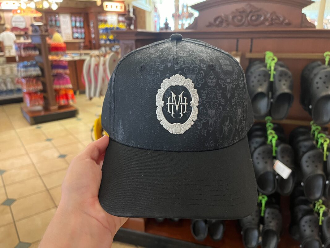 A person holds a black baseball cap with an ornate "HM" logo. In the background, a store display features various items, including Crocs and souvenirs.
