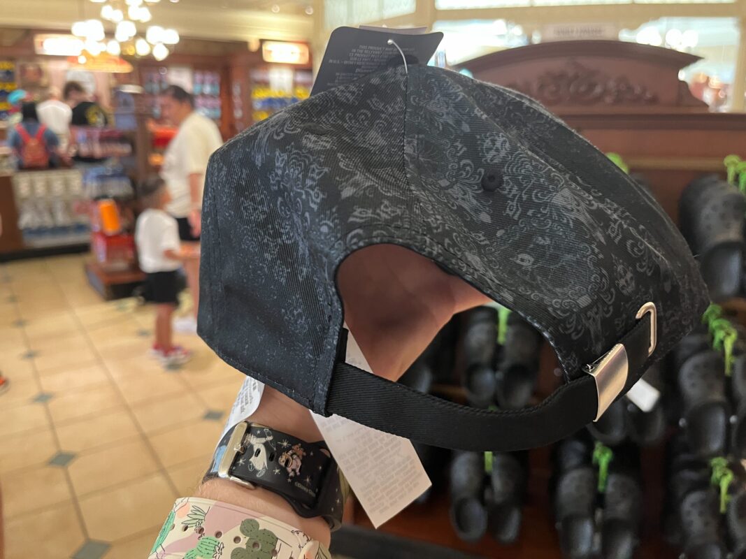 A person holding a black patterned cap by its adjustable strap inside a store. In the background, there are shelves with various items and a few people shopping.