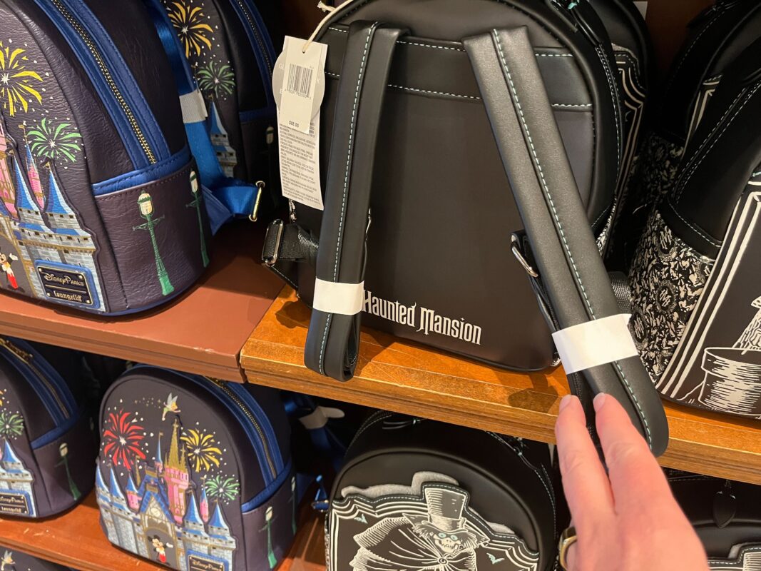 Several themed backpacks displayed on shelves in a store. A hand is visible, seemingly picking up a black backpack labeled "Haunted Mansion." Other backpacks feature various designs, including a castle.