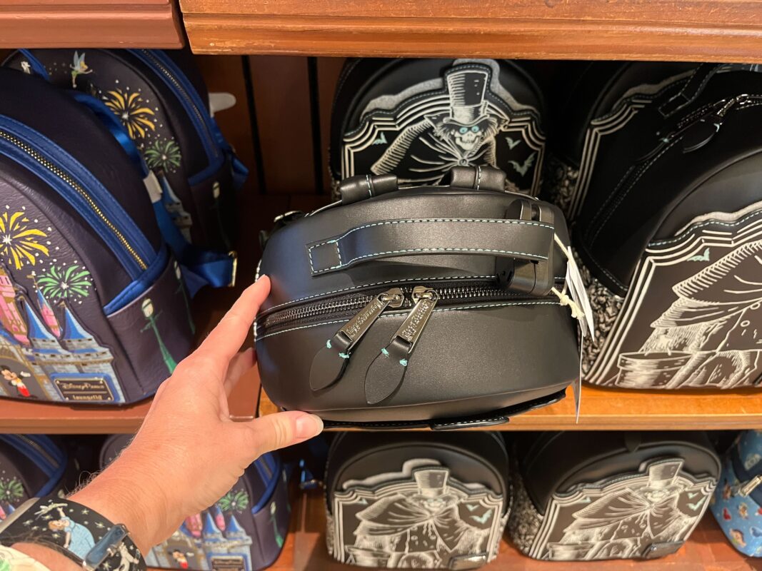 A person holds a black backpack with silver zippers in front of a display shelf containing other similar backpacks with various designs.