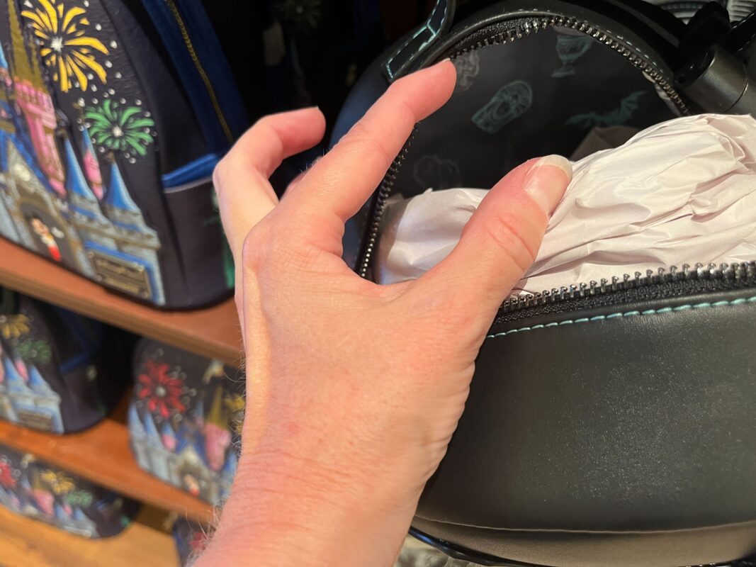 A hand opens a black rounded zipper bag, revealing crumpled tissue paper inside. In the background, various bags with colorful designs are displayed on shelves.