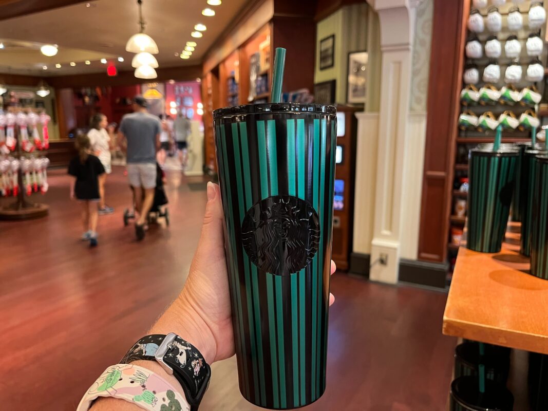A hand holds a striped green Starbucks tumbler with a straw inside a well-lit store. Other people and store items are visible in the background.
